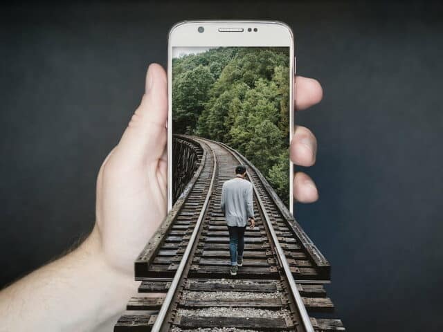 Main tenant un téléphone. On y voit une photo de rails au milieu des arbres avec un homme marchant au centre. La photo sort de l'écran et les rails arrivent au premier plan