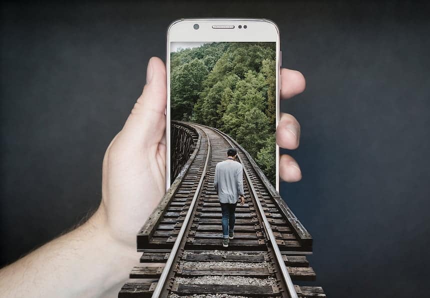 Main tenant un téléphone. On y voit une photo de rails au milieu des arbres avec un homme marchant au centre. La photo sort de l'écran et les rails arrivent au premier plan