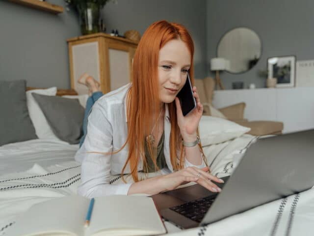 Femme rousse allongée sur le ventre sur son lit, devant un ordinateur, un téléphone portable à l'oreille