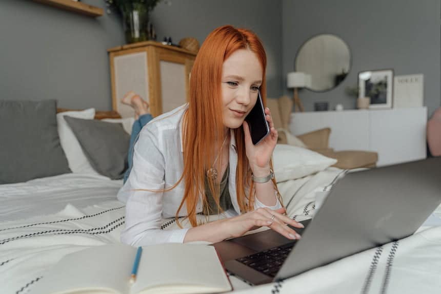 Femme rousse allongée sur le ventre sur son lit, devant un ordinateur, un téléphone portable à l'oreille