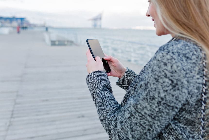 Portrait de belle jeune femme souriante, en manteau gris utilisant son smartphone près de la mer