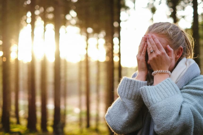 Femme dans une forêt en journée, portant un manteau gris avec les mains sur son visage 